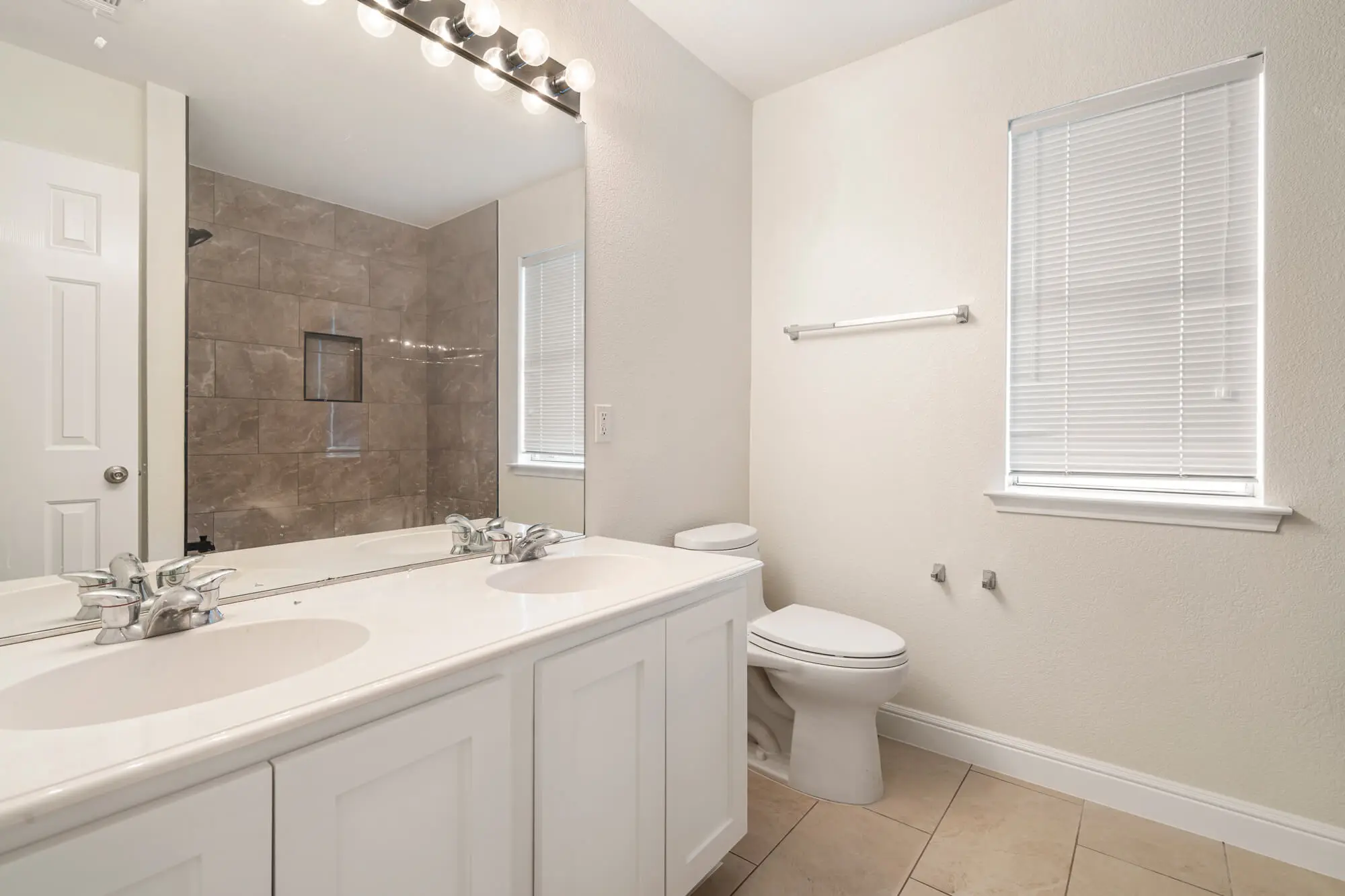 Double sink bathroom with freshly painted cream walls, white shaker vanity cabinet, white baseboards, and tile shower surround visible in mirror on a DFW interior painting project by J&R Alliance Painting LLC