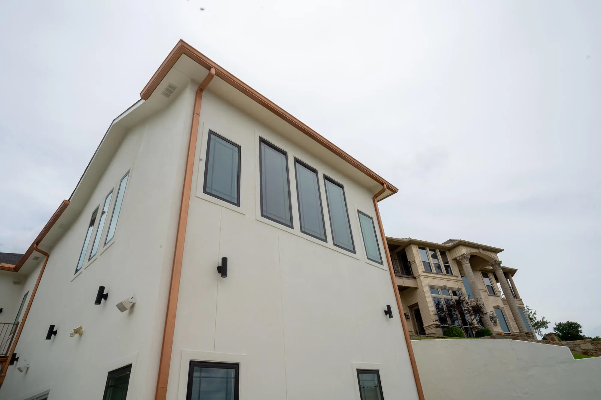 Angled upper elevation view of a completed white stucco exterior repaint with copper gutters and charcoal window trim on a DFW estate home with a neighboring luxury property in the background