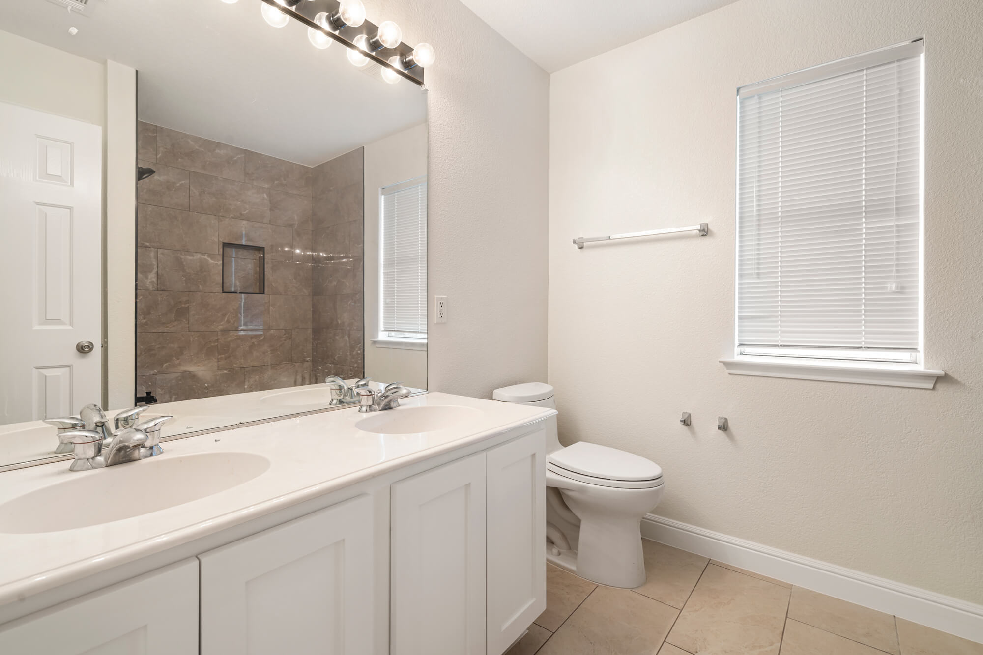Double sink bathroom with freshly painted cream walls, white shaker vanity cabinet, white baseboards, and tile shower surround visible in mirror on a DFW interior painting project by J&R Alliance Painting LLC